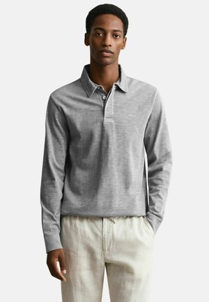 Man draagt een lichtgrijs poloshirt met lange mouwen en kraag, en beige broek met trekkoord, staat met één hand in de zak.