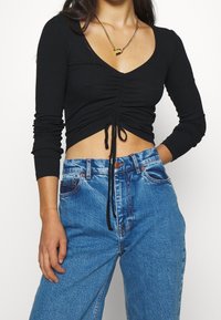 Schwarzes geripptes Langarmtop mit Zugbanddetails und V-Ausschnitt, kombiniert mit hellblauen hoch taillierten Jeans.