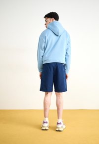 Svijetloplava hoodie s vezicom i rebrastim manžetama, u kombinaciji s mornarsko plavim šortsem. Model nosi bijele tenisice s ljubičastim detaljima, stoji na žutom tepihu.