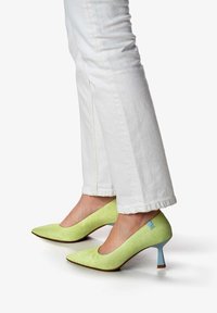 Chaussures à talons aiguilles en satin vert avec un bout pointu et des accents bleu clair, associées à un pantalon en denim blanc effiloché.