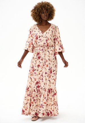 Kobieta o kręconych włosach, ubrana w jasnoróżową, kwiecistą sukienkę maxi z rękawami do łokcia i marszczoną talią, stojąca na białym tle.