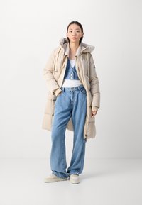 Beige gewatteerde jas met een capuchon, gedragen over een witte tanktop en blauwe denim salopette. Loszittende jeans en lichtgekleurde sneakers maken de look af.
