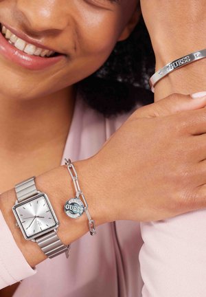 Zilveren horloge met een vierkante wijzerplaat en een metalen band, gecombineerd met een zilveren kettingarmband met een "GUESS" hangertje en kleine heldere accenten.