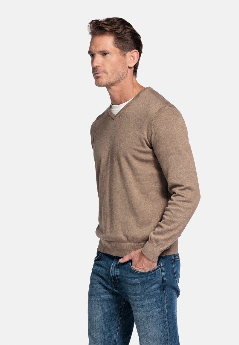 Tan v-hals trui gemaakt van zachte stof, gecombineerd met blauwe denim jeans. De trui heeft een geribde zoom en boorden, wat zorgt voor een aansluitende look.