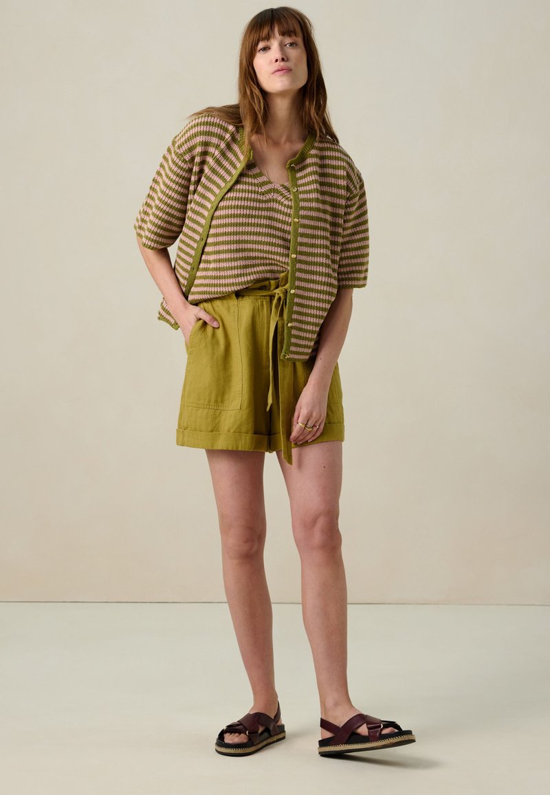 Veste en maille légère à rayures beige et olive, associée à un short à cordon de serrage olive et des sandales marron foncé à semelle épaisse.