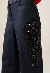 Pantalones de mezclilla oscura con un intrincado diseño de lentejuelas negras con motivos florales en el muslo izquierdo. La tela tiene una textura suave y un ajuste estructurado.