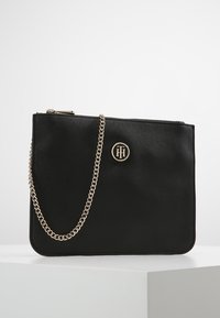 Bolso sintético negro con cierre de cremallera, correa de cadena dorada y un logo redondo en la parte frontal. Textura suave y forma rectangular.