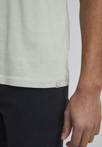 Tricou scurt cu mânecă, verde deschis, dintr-un material texturat. Prezintă o etichetă mică cu logo la tiv și un design subtil aproape de cusătura laterală.