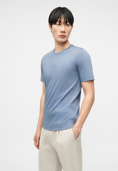 Νέος άνδρας όρθιος με τα χέρια στις τσέπες, φορούν ανοιχτό μπλε κοντομάνικο μπλουζάκι και ανοιχτό μπεζ παντελόνι, μπροστά σε απλό φόντο.