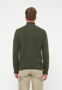 Muž v tmavozelenom pletenom svetri a béžových cargo nohaviciach, zobrazený zozadu na jednoduchom bielom pozadí.
