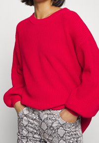 Fuchsia striktrøje med en løs pasform og ribbet tekstur, sammen med tætsiddende grå slangeskindsbukser, der viser en bred udskæring og opsmøgede ærmer.