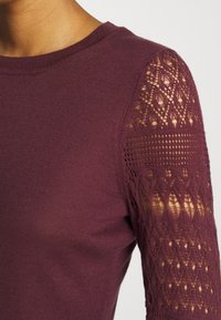 Burgunderfarbener Langarmshirt mit gestricktem Muster an den Ärmeln, weicher Textur und klassischem Rundhalsausschnitt.