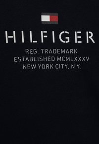 Granatowy bawełniany t-shirt z logo "HILFIGER" w białym nadruku, z detalami zarejestrowanego znaku towarowego oraz małym emblematem flagi powyżej.