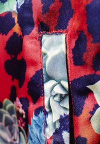 Květinová tkanina s živými červenými, modrými a zelenými barvami a hladkou strukturou. Obsahuje kontrastní panel s velkými vzory listů.