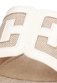 Chanclas blancas que presentan un gran logo en relieve sobre una banda de textil tejido en color beige. Plantilla suave y acolchada con textura en marrón claro.