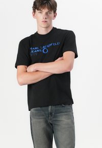 Svart bomull t-skjorte med blå grafisk tekst som leser "KARL LAGERFELD JEANS" over brystet. Modell iført lyseblå denimjeans.