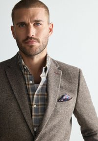 Brown herringbone blazer paired with a light blue plaid shirt. Features a colorful pocket square and textured fabric.
