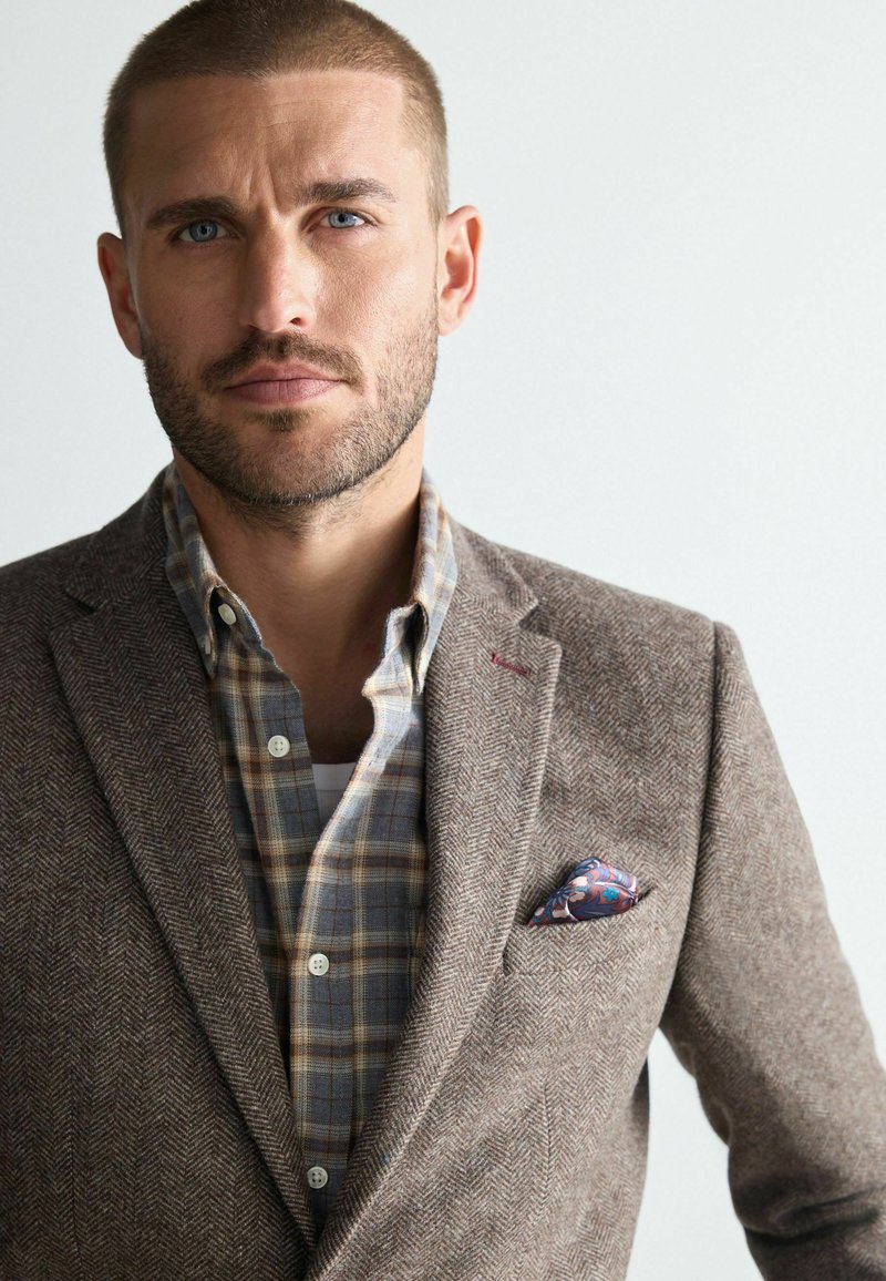 Brown herringbone blazer paired with a light blue plaid shirt. Features a colorful pocket square and textured fabric.