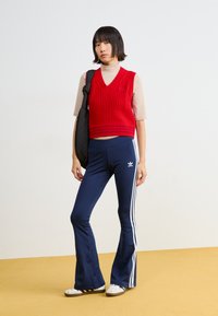 Piros kötött mellény V nyakkal, sötétkék, bővülő sportnadrággal, fehér oldalsávokkal és egy kis logóval párosítva. Fehér sportcipő.