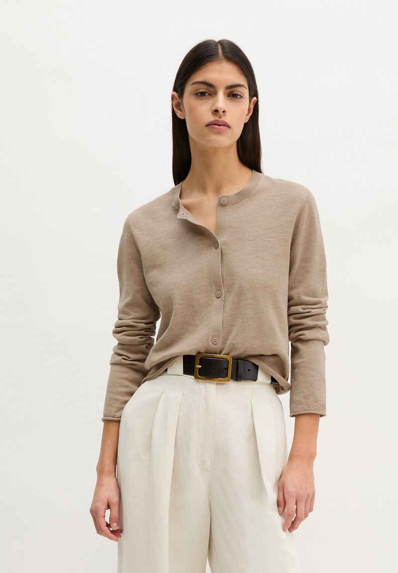 Femme portant un cardigan beige boutonné rentré dans un pantalon blanc plissé taille haute, accompagné d'une ceinture noire avec une boucle dorée.