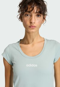 Vrouw met krullend haar die een lichtblauw Adidas T-shirt met wit logo draagt, tegen een effen achtergrond, met een neutrale gezichtsuitdrukking vooruitkijkend.