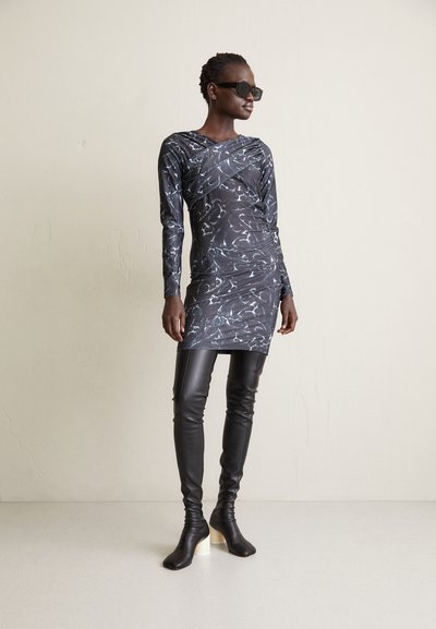 Čierne šaty s dlhým rukávom a bielym abstraktným vzorom, skombinované s tesnými čiernymi koženými nohavicami a so zúženými kotníkovými topánkami na podpätku.