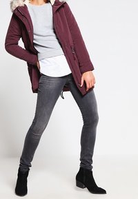 Burgunderröd vadderad jacka med pälsfodrad huva, grå tröja, vit skjorta och grå slim-fit jeans. Svarta ankelskånsar fulländar outfiten.