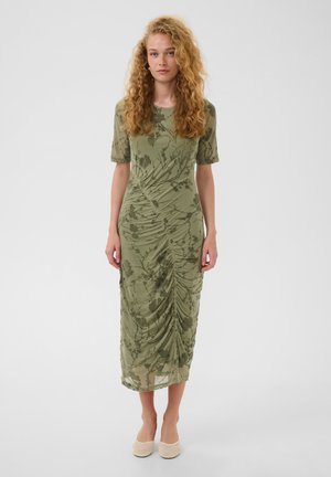 Femme aux cheveux bouclés portant une robe midi verte ajustée avec des détails froncés et des motifs inspirés de la nature, associée à des ballerines beiges tissées.