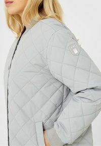 Kobieta w jasnoszarej kurtce bombowcu z pikowaniem, z ściągaczami w mankietach i małą naszywką z logo na górnej części rękawa.