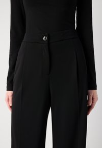 Pantaloni neri su misura con una cintura elegante e un bottone argentato, profonde pieghe e tasche laterali, realizzati in un tessuto liscio.