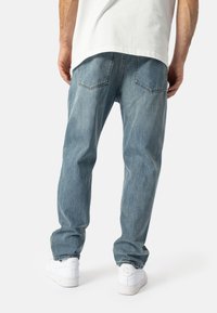 Uomo che indossa jeans azzurri chiari, sneakers bianche e una camicia bianca, mostrato di spalle su uno sfondo chiaro e semplice.