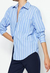 Ljusblå skjorta med knappar och vita vertikala ränder, bröstficka, klassisk krage och uppkavlade ärmar, tillverkad av mjukt tyg.
