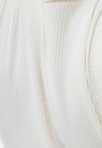 Bílý žebrovaný top bez rukávů s detailním pohledem na texturovanou látku a hladkým výstřihem. Design zdůrazňuje vertikální linie a přiléhavý střih.