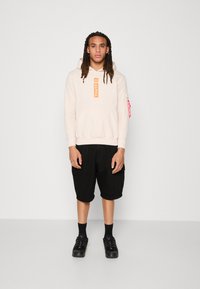 Felpa con cappuccio beige con logo arancione "ALPHA", abbinata a pantaloni cargo neri e sneakers nere. Presenta tasca a marsupio e dettagli con zip laterale.