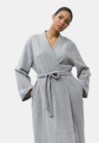 Accappatoio grigio in tessuto texture, con maniche ampie, vita con cinturino e due tasche laterali. Design semplice e funzionale.