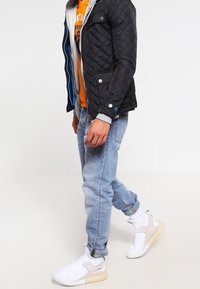Sort quiltet jakke med grå hætteforing, blå lynlåsdetaljer, orange grafisk t-shirt, lyseblå jeans og hvide sneakers.