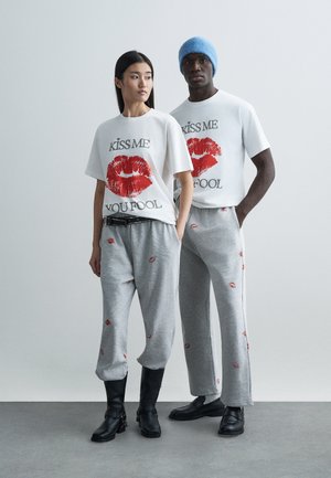 Λευκά γραφικά t-shirts με κόκκινη εκτύπωση χειλιών και μαύρο κείμενο, συνδυασμένα με γκρι παντελόνια φόρμας με κόκκινα σχέδια φιλιά· μαύρα και γκρι παπούτσια.