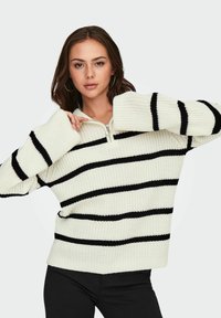 Pull en tricot épais crème avec des rayures horizontales noires, doté d'un col zippé mi-haut, de larges manches et d'un tissu texturé.