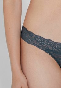 Tanga en dentelle dans un bleu-vert foncé avec un motif floral, une texture douce et une taille délicate à bordure festonnée pour un design minimaliste.
