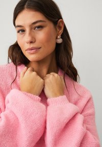 Maglione rosa chiaro lavorato a maglia con spalle abbassate e polsini a costine, accessoriato con orecchini rotondi beige e oro. Texture liscia.