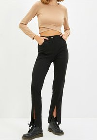 Schwarze, taillierte Hosen mit gerader Form, die seitliche Schlitze am Saum aufweisen. Kombiniert mit einem beigen Langarm-Crop-Top und schwarzen Stiefeln.
