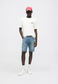 Λευκό βαμβακερό T-shirt με γραφικό σχέδιο, συνδυασμένο με μπλε τζιν σορτς. Κόκκινο καπέλο μπέιζμπολ, λευκά αθλητικά παπούτσια και λευκές κάλτσες ολοκληρώνουν την εμφάνιση.