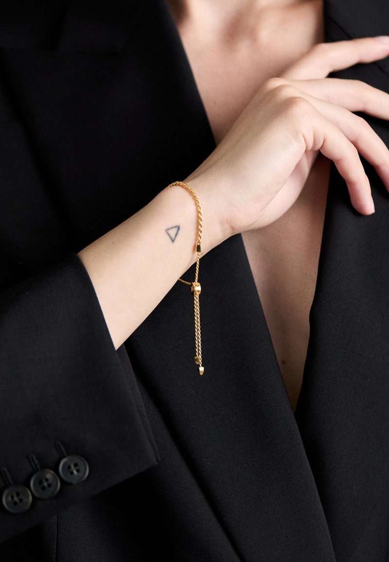 Kuldne kettkäevõru tekstuuriga viimistlusega ja reguleeritava lukkudega, kantud randmel, millel on must kolmnurkne tätoveering, tumeda bleiseri taustal.