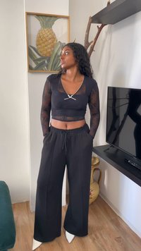 Mulher em top cropped de renda preta e calças largas pretas, de pé no interior perto de uma TV e uma obra de arte emoldurada de abacaxi, olhando de lado com as mãos nos bolsos.