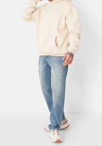 Beige Kapuzenpullover mit einer Fronttasche, kombiniert mit hellblauen Jeans und cremefarbenen Sportschuhen mit orangefarbenen Akzenten.