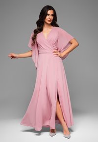 En rosa chiffongklänning med omslagdesign, fladdrande ärmar och sidoklyfta. Tyget har en mjuk struktur och en flytande fall, kompletterad av glittriga klackar.