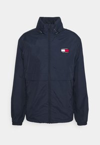 Chaqueta cortavientos navy con cuello alto, cierre completo y puños elásticos. Presenta un pequeño logo en rojo y blanco en el pecho. Textura suave.
