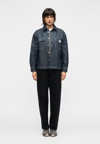 Τζιν μπουφάν με δύο τσέπες στο στήθος, σκούρο μπλε χρώμα και λευκή ραφή, φορεμένο πάνω από ριγέ πουκάμισο και μαύρο παντελόνι. Γκρι αθλητικά παπούτσια ολοκληρώνουν την εμφάνιση.