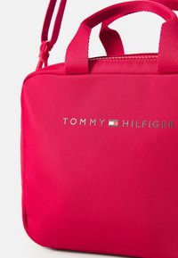 Sac en tissu rose vif avec poignées supérieures doubles et une bandoulière ajustable. Présente un logo argenté "TOMMY HILFIGER" et une fermeture éclair.