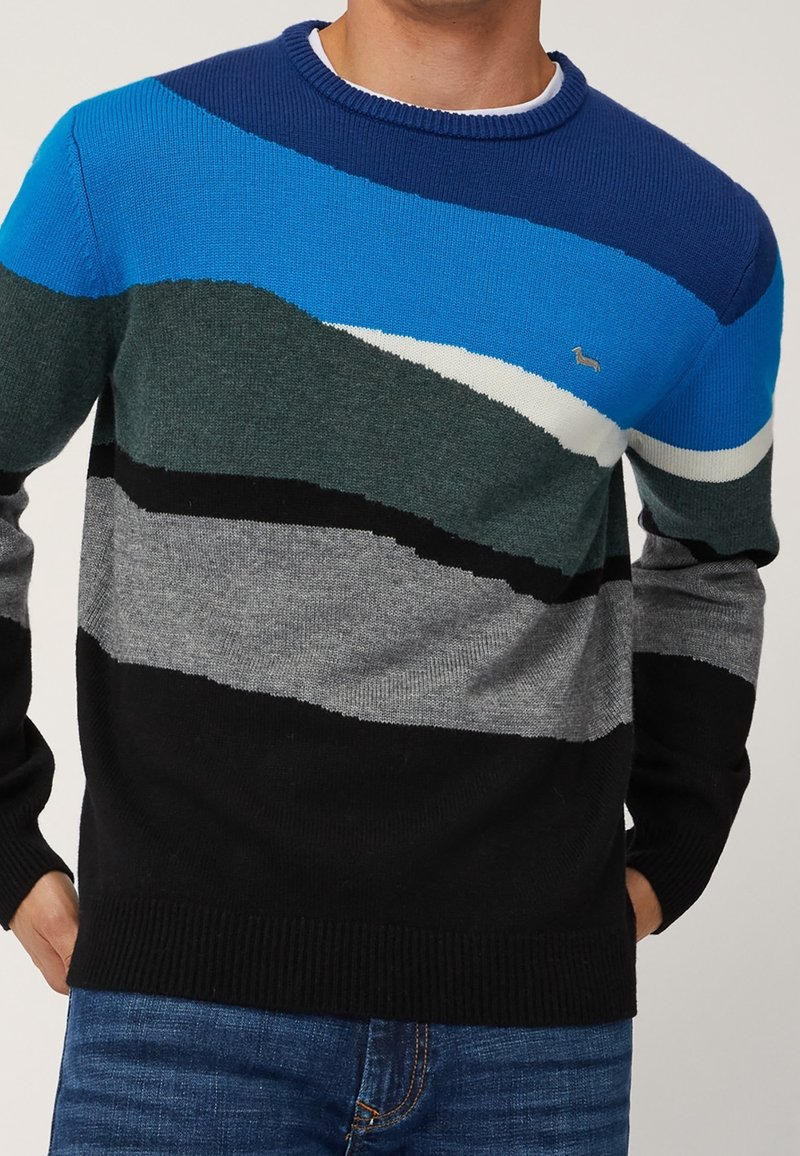 Homme portant un pull à rayures multicolores en bleu, vert, gris, noir et blanc avec un petit logo sur la poitrine, associé à un jean bleu.